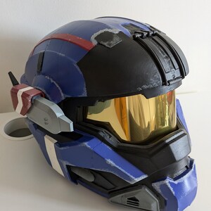 Pode incluir: Um capacete azul e preto com viseira dourada. O capacete tem detalhes vermelhos e brancos e uma superf&iacute;cie texturizada. Possui um dispositivo de comunica&ccedil;&atilde;o lateral. Provavelmente &eacute; um item de cosplay ou um adere&ccedil;o.