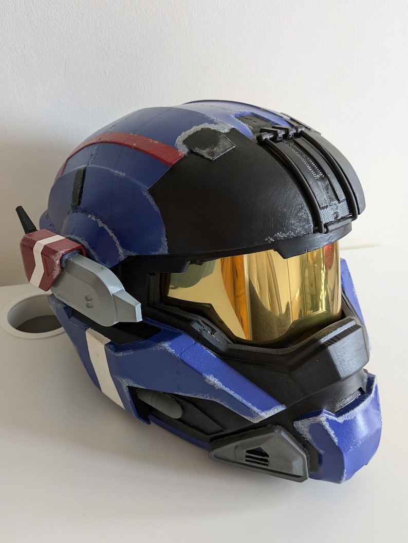 Pode incluir: Um capacete azul e preto com viseira dourada. O capacete tem detalhes vermelhos e brancos e uma superf&iacute;cie texturizada. Possui um dispositivo de comunica&ccedil;&atilde;o lateral. Provavelmente &eacute; um item de cosplay ou um adere&ccedil;o.