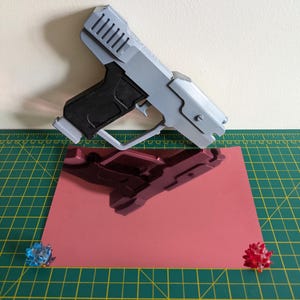 Pode incluir: Uma pistola de brinquedo cinza e preta com um design detalhado, repousando sobre uma superf&iacute;cie rosa reflexiva. A pistola est&aacute; angulada, mostrando sua forma. Dois pequenos objetos decorativos coloridos s&atilde;o colocados perto das bordas da superf&iacute;cie rosa. O fundo &eacute; bege claro.