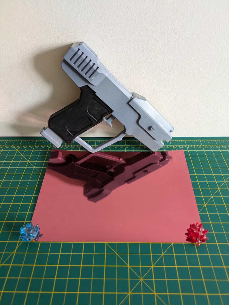 Pode incluir: Uma pistola de brinquedo cinza e preta com um design detalhado, repousando sobre uma superf&iacute;cie rosa reflexiva. A pistola est&aacute; angulada, mostrando sua forma. Dois pequenos objetos decorativos coloridos s&atilde;o colocados perto das bordas da superf&iacute;cie rosa. O fundo &eacute; bege claro.
