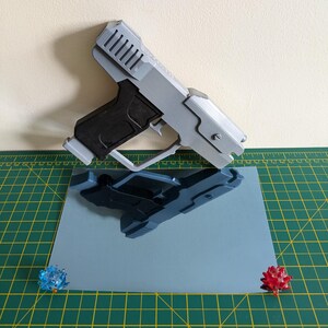 Pode incluir: Uma pistola de brinquedo cinza e preta com um design detalhado, repousando sobre uma superf&iacute;cie azul clara. O reflexo da pistola &eacute; vis&iacute;vel. Dois pequenos objetos decorativos, coloridos e pontiagudos s&atilde;o colocados nos cantos da superf&iacute;cie azul.