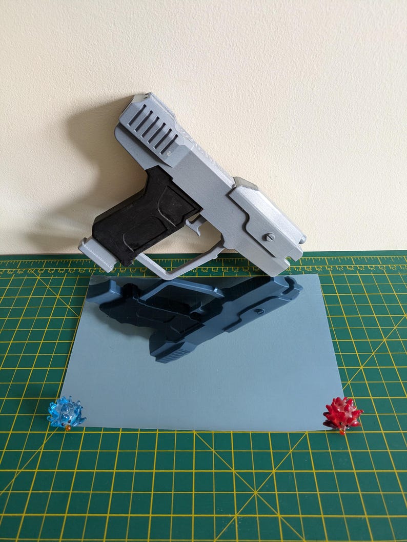 Pode incluir: Uma pistola de brinquedo cinza e preta com um design detalhado, repousando sobre uma superf&iacute;cie azul clara. O reflexo da pistola &eacute; vis&iacute;vel. Dois pequenos objetos decorativos, coloridos e pontiagudos s&atilde;o colocados nos cantos da superf&iacute;cie azul.