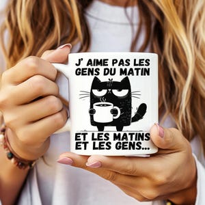 Peut inclure: Mug en céramique blanche avec un chat noir grincheux tenant une tasse de café. Le mug porte l'inscription en français : "J'aime pas les gens du matin et les matins et les gens..."