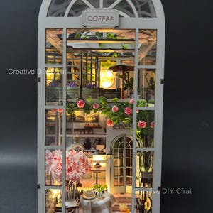May include: A miniature, illuminated coffee shop model with a white frame and arched window. The model features a detailed interior with tables, chairs, and floral arrangements. The sign above the window reads "COFFEE".