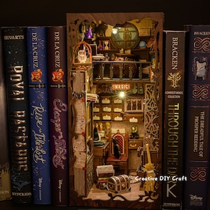 May include: A miniature bookshelf diorama with a magical apothecary shop inside. The shop has a wooden counter, shelves filled with potion bottles, and a black cat sitting in a chair. The diorama is made of wood and is painted in a dark brown color. The diorama is displayed on a shelf with other books.