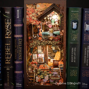 May include: A 3D wooden model of a small cabin with a lit window, surrounded by autumn leaves and a welcome sign. The model is displayed on a bookshelf with other books.