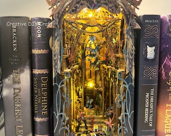 Puede incluir: Un separador de libros de madera con forma de castillo en miniatura con un interior brillante. El separador de libros está flanqueado por cinco novelas de fantasía con títulos como "The Darkest Minds" y "Delphine and the Silver Needle".