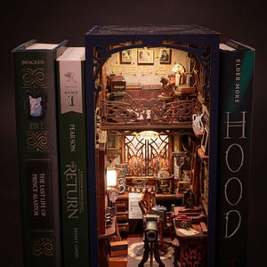 May include: A miniature room diorama built inside a book. The room is lit with a lamp and features a desk, a chair, and a camera on a tripod. The book is titled "The Return" and is part of a series of books titled "A Kingdom Kalplay Novel".