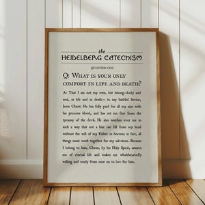 May include: A framed print featuring text from the Heidelberg Catechism. The text is in black on a cream-colored background, with a wooden frame. The print is displayed against a white wall and wooden floor.