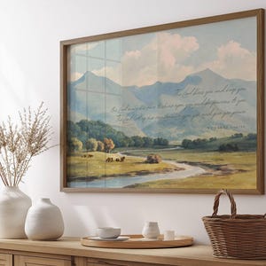 May include: A framed landscape print featuring a serene valley scene with mountains, a river, and text. The artwork is in a wooden frame and is displayed on a wooden cabinet. Decorative vases and a woven basket are also visible.