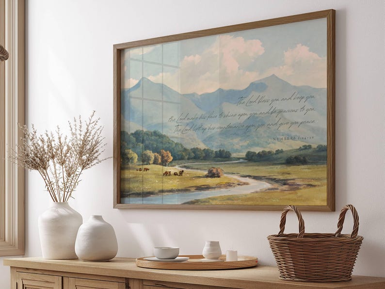 May include: A framed landscape print featuring a serene valley scene with mountains, a river, and text. The artwork is in a wooden frame and is displayed on a wooden cabinet. Decorative vases and a woven basket are also visible.