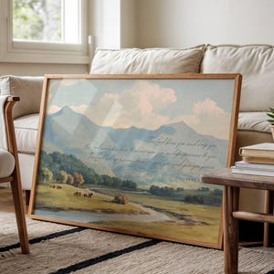 May include: A framed landscape print featuring a serene mountain scene with a river and text. The artwork is in a wooden frame and leans against a beige sofa. A small succulent in a pot sits on a side table.