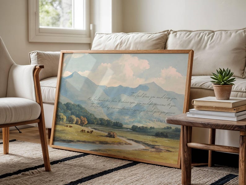 May include: A framed landscape print featuring a serene mountain scene with a river and text. The artwork is in a wooden frame and leans against a beige sofa. A small succulent in a pot sits on a side table.