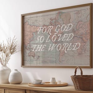 May include: A framed world map print with the text "FOR GOD SO LOVED THE WORLD" in white. The map is in muted tones of pink, blue, and beige. A wooden basket and vases are on a wooden surface.