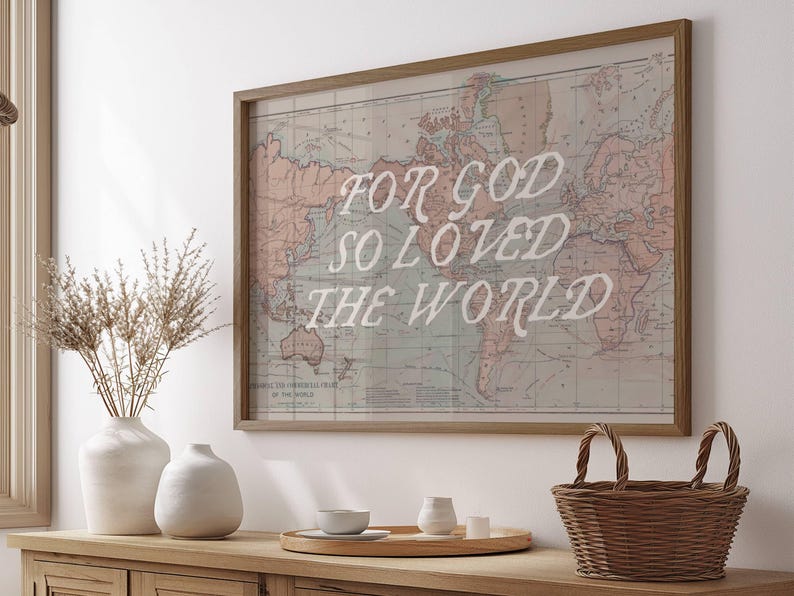 May include: A framed world map print with the text "FOR GOD SO LOVED THE WORLD" in white. The map is in muted tones of pink, blue, and beige. A wooden basket and vases are on a wooden surface.