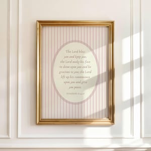 Op de afbeelding: Een ingelijste print met een goudkleurige lijst. Het kunstwerk heeft een lichtroze en wit gestreepte achtergrond met een ovale tekstvak. De tekst luidt "The Lord bless you and keep you..." uit Numeri 6:24-26.