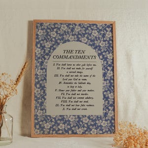 Puede incluir: Una impresión enmarcada con "LOS DIEZ MANDAMIENTOS" sobre un fondo floral azul. El texto está centrado en un fondo gris claro. Un jarrón de cristal transparente con flores secas está en primer plano.
