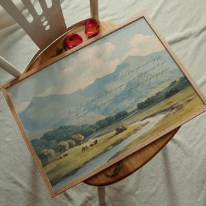 May include: A framed landscape print featuring a pastoral scene with mountains, a river, and trees. The artwork has a light wood frame and includes script text. Two red rose petals rest on the chair.