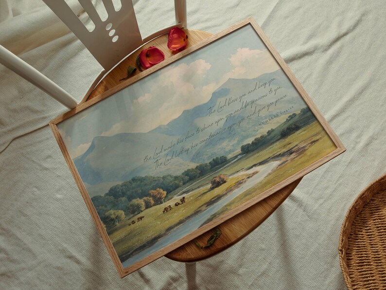 May include: A framed landscape print featuring a pastoral scene with mountains, a river, and trees. The artwork has a light wood frame and includes script text. Two red rose petals rest on the chair.