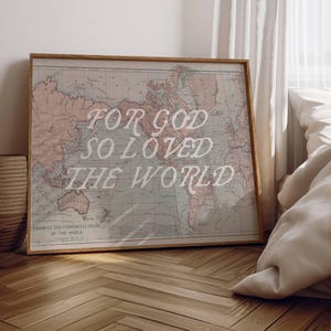 May include: Framed vintage-style world map with the text "FOR GOD SO LOVED THE WORLD" in white script. The map has a light pink and blue color scheme and is displayed on a wooden floor. A woven basket and a bed are in the background.