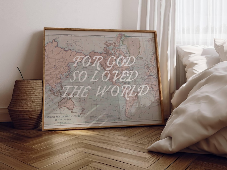 May include: Framed vintage-style world map with the text "FOR GOD SO LOVED THE WORLD" in white script. The map has a light pink and blue color scheme and is displayed on a wooden floor. A woven basket and a bed are in the background.