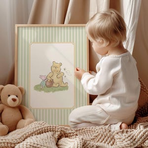 May include: A framed illustration of Winnie the Pooh and Piglet, set against a light green and white striped backdrop. A soft teddy bear rests on a beige blanket. A child in white clothing is looking at the framed picture.