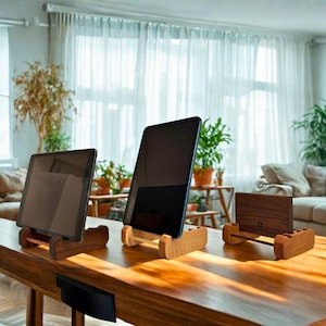 May include: Three wooden tablet stands in varying designs. The stands are made of light and dark wood, with tablets displayed on them. The stands are on a wooden table in a well-lit room with plants and a window in the background.