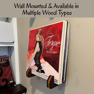 May include: A wall-mounted record holder made of dark wood, displaying a stack of vinyl records. The holder is mounted on a light gray wall. The album cover visible features Fergie, with the text "Fergie THE DUTCHESS". The text "Wall Mounted & Available in Multiple Wood Types" is also visible.