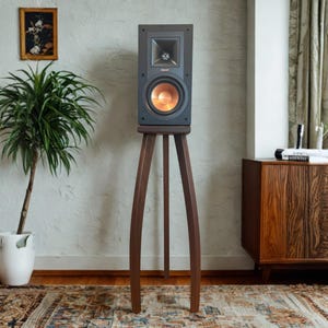 May include: A dark gray Klipsch bookshelf speaker with a prominent metallic copper-colored woofer and a square horn tweeter, mounted on a tall, three-legged wooden tripod stand with curved legs that taper toward the floor.