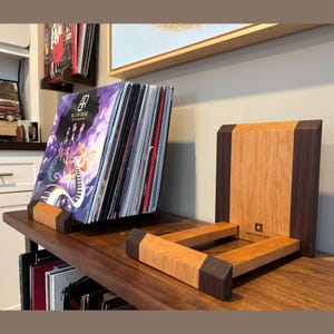 May include: Wooden record holder with a cherry and walnut design. The holder is designed to display vinyl records. The holder is on a dark wooden surface. The record holder is made by Greene Wood Design.