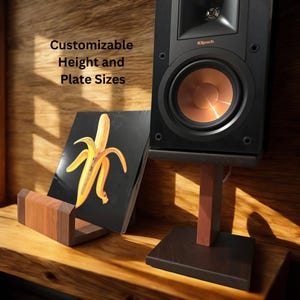 May include: A black speaker with a copper-colored cone sits on a dark wood stand. A wooden record holder displays a record with a banana graphic. The text "Customizable Height and Plate Sizes" is visible.