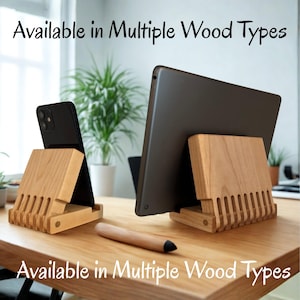May include: Wooden phone and tablet stands with a slotted design, holding a black smartphone and a tablet. A wooden stylus rests on the table. The text "Available in Multiple Wood Types" is displayed. The stands are made of light-colored wood.
