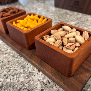 May include: A wooden serving tray with three small, square wooden bowls. The bowls are filled with almonds, orange snacks, and pistachios. The tray and bowls are made of a reddish-brown wood.