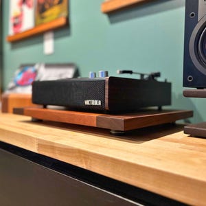 May include: A black Victrola record player with wooden accents sits atop a two-tiered wooden stand. The stand is made of a lighter wood with darker wood corner accents. The record player has silver knobs and a mesh speaker cover.