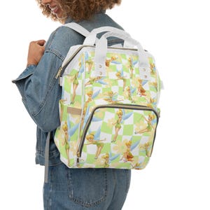 Puede incluir: Una mochila blanca y verde con un patrón de tablero de ajedrez con un hada de dibujos animados. La mochila tiene un asa superior, correas para los hombros y múltiples bolsillos. El hada lleva un vestido verde y tiene alas azules.
