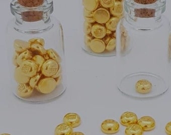May include: Three small glass bottles with cork stoppers filled with gold-colored beads. The bottles are on a white surface with several gold-colored beads scattered around them.