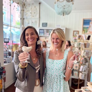 May include: Two women smiling and holding up small glasses with white, decorative items inside. The woman on the left is wearing a brown leather jacket, a white skirt, and gold jewelry. The woman on the right is wearing a green floral dress and a gold necklace.