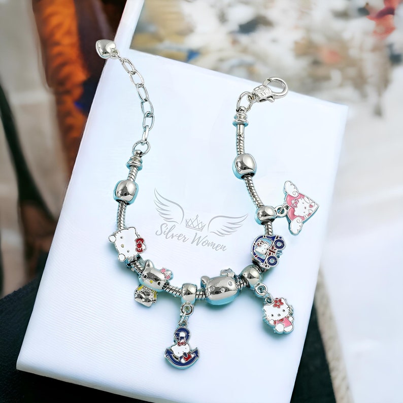May include: A silver charm bracelet with multiple charms, including a Hello Kitty charm, a car charm, an anchor charm, and a heart charm. The bracelet is on a white background.