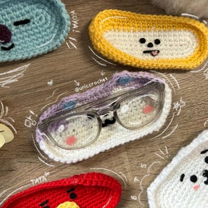 May include: A selection of handmade crochet eyeglass cases in various colours and designs. One white case with a purple top holds eyeglasses. Other cases are blue, yellow, and red, each with a unique cartoon face. The cases are on a wooden surface.
