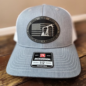 May include: Gray and white trucker hat with the text "DRILL BABY DRILL" and an image of an American flag and oil derrick. The hat has a Richardson patch that reads "ORIGINAL 112 TRUCKER ADJUSTABLE".