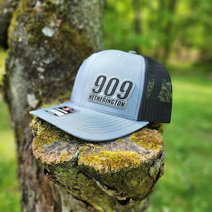 May include: A gray and black baseball cap with a black mesh back. The front of the cap has a silver patch with the number "909" and the text "Hetherington" in black.