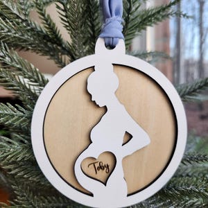 May include: White wooden ornament shaped like a pregnant woman with a heart cutout in the belly. The heart has the name "Toby" engraved inside. The ornament is hanging on a blue ribbon on a Christmas tree.