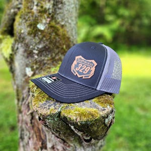 May include: A black and gray trucker hat with a brown leather patch that says "DiPrizio 329". The hat is sitting on a tree stump.