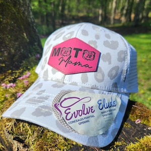 May include: A white trucker hat with a gray leopard print pattern. The hat features a pink patch with the words "Moto Mama" and a concealed ponytail opening. The hat also has a makeup-resistant sweatband.