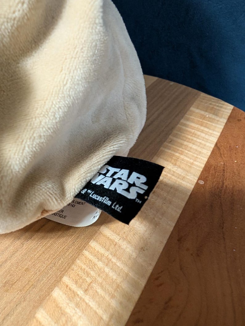 Puede incluir: Un juguete de peluche blanco con una etiqueta negra que dice "STAR WARS" y "& Lucasfilm Ltd."