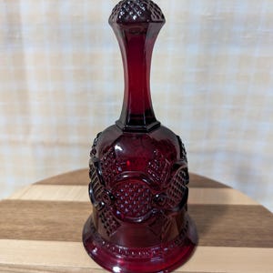 May include: A red glass bell with a textured surface and a diamond-shaped top. The bell is sitting on a wooden surface.