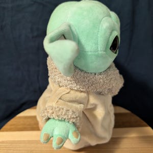 Puede incluir: Un juguete de peluche del personaje Baby Yoda de la serie de Star Wars The Mandalorian. El juguete lleva un abrigo marr&oacute;n y blanco con un cuello de piel. El juguete es verde y tiene ojos grandes.