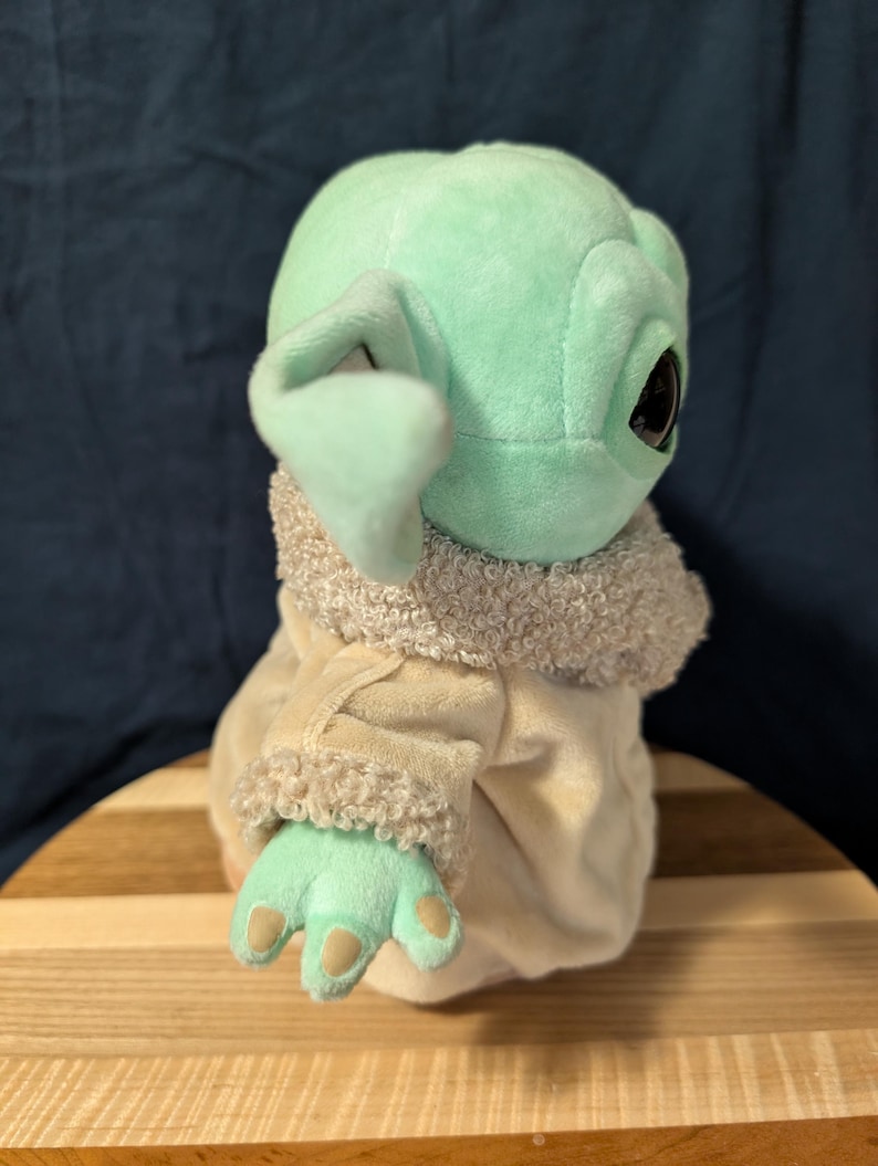 Puede incluir: Un juguete de peluche del personaje Baby Yoda de la serie de Star Wars The Mandalorian. El juguete lleva un abrigo marr&oacute;n y blanco con un cuello de piel. El juguete es verde y tiene ojos grandes.