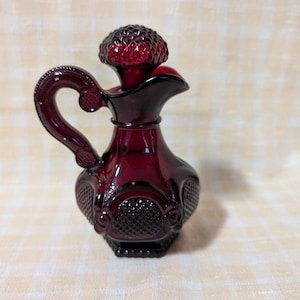 May include: A ruby red glass pitcher with a handle and a decorative stopper. The pitcher has a textured pattern and a spout for pouring. The stopper is a rounded shape with a textured surface. The pitcher is likely for serving beverages.
