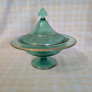May include: A translucent green glass candy dish with a matching lid featuring a fleur-de-lis finial. The bowl and lid are accented with a thin gold rim and an embossed floral pattern around the center. The dish stands on a circular pedestal base.